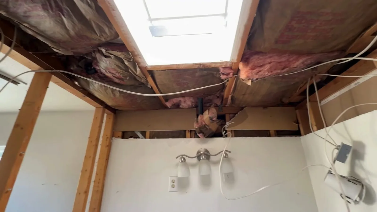 Bathroom ceiling opened up to show wooden joists, pink insulation and wiring above a vanity light