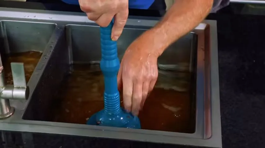 Hand pressing a blue plunger down in a stainless steel sink filled with brown, murky water