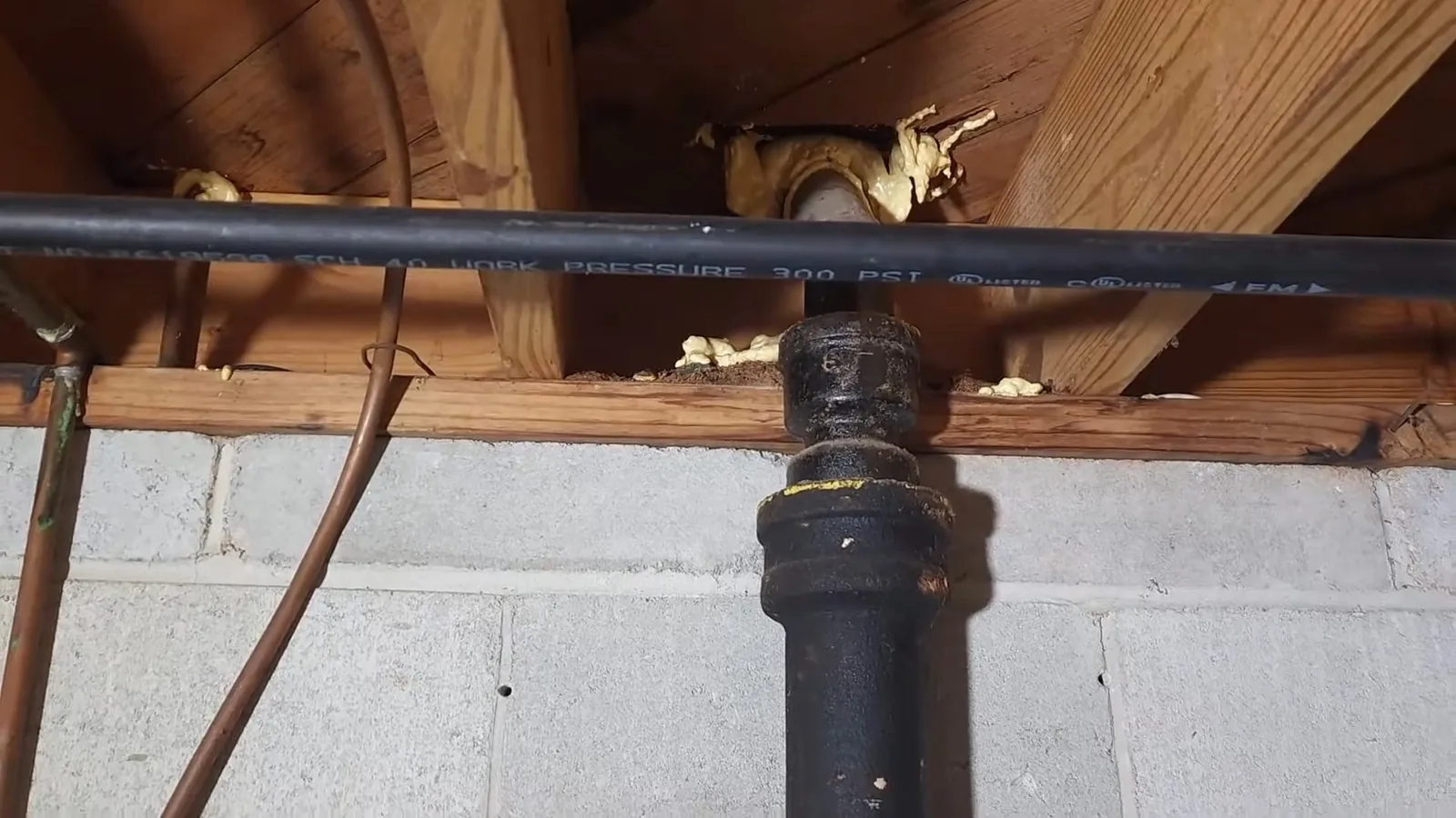 Black cast iron drain fitting rising through a basement floor joist beside a black plastic pipe and a copper line, with a concrete block wall behind