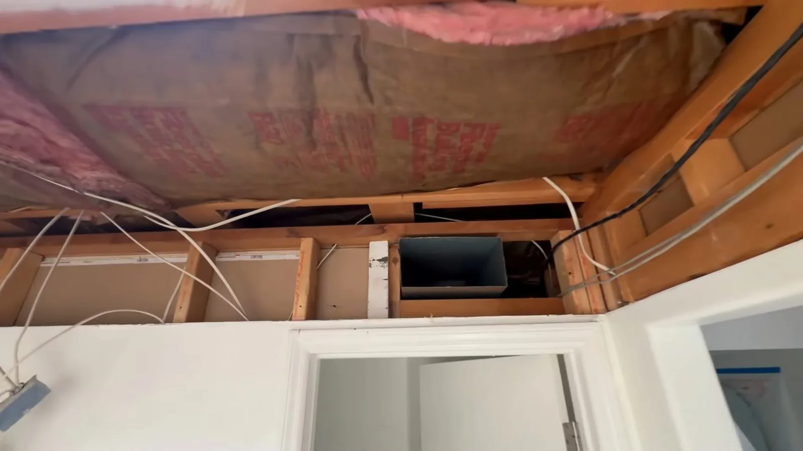 Opened ceiling above a doorway showing wooden framing, pink insulation and a metal duct box