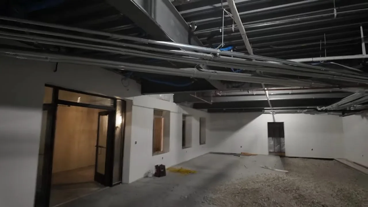 Large empty commercial space under construction with an open ceiling full of pipes and conduit, glass doors at the left and a gravel floor