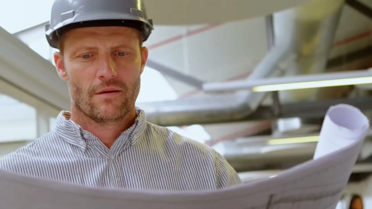 Man in a white hard hat and striped shirt studying a roll of building plans, with pipes and ductwork behind him
