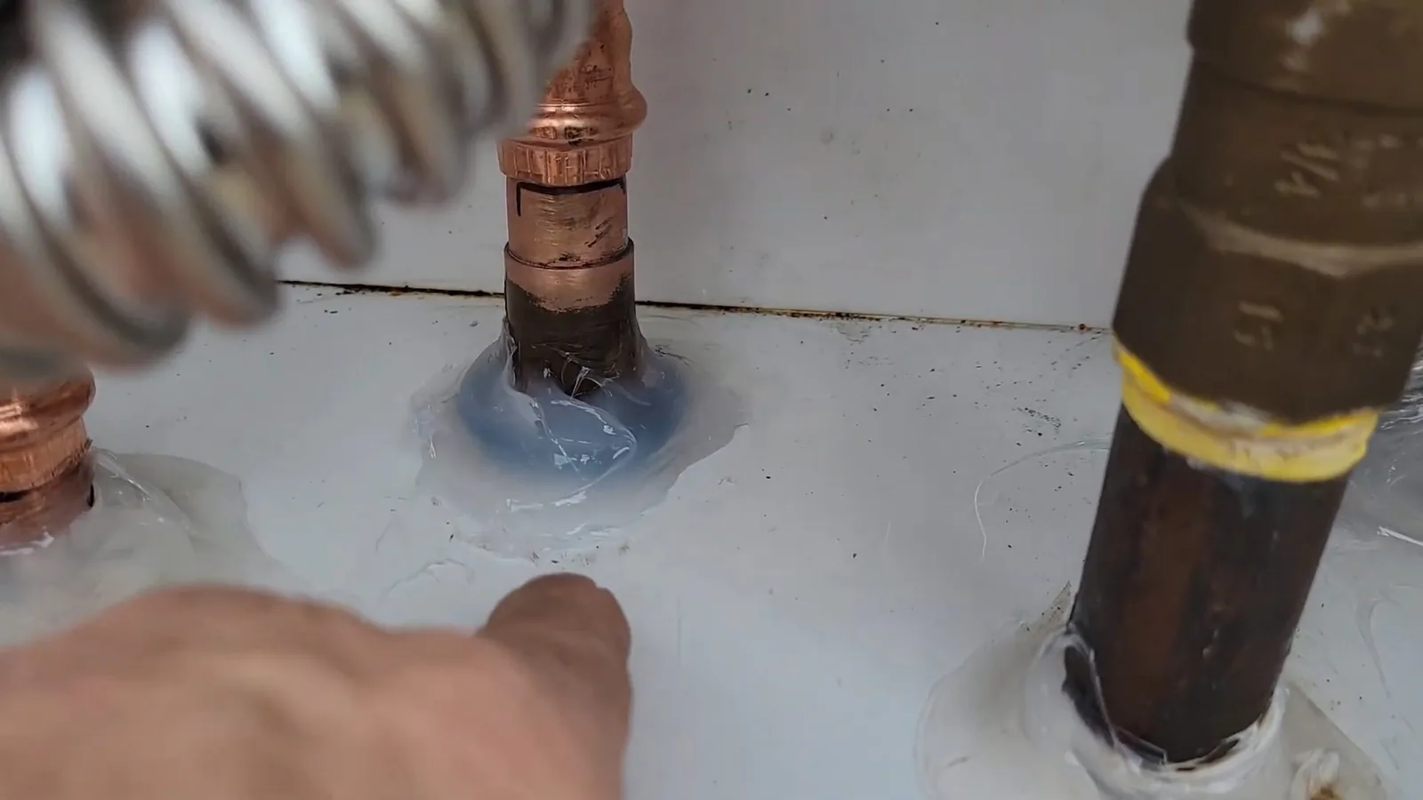 Top of a white water heater with copper and brass pipes coming out of it, a coiled metal hose at upper left and a finger pointing at one fitting