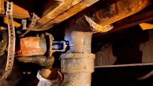Heavily rusted cast iron drain fitting under wooden floor framing, with a perforated metal strap and a copper fitting beside it