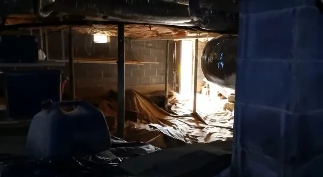 Dim crawl space with black plastic sheeting on the ground, a block pier at right and a blue jug beside the sheet