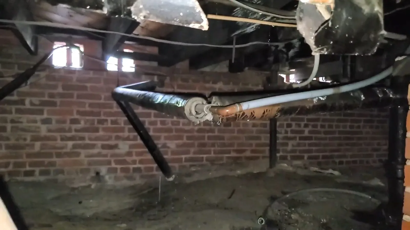 Crawl space with a brick foundation wall, a dirt floor and old black and silver pipes running along the floor framing