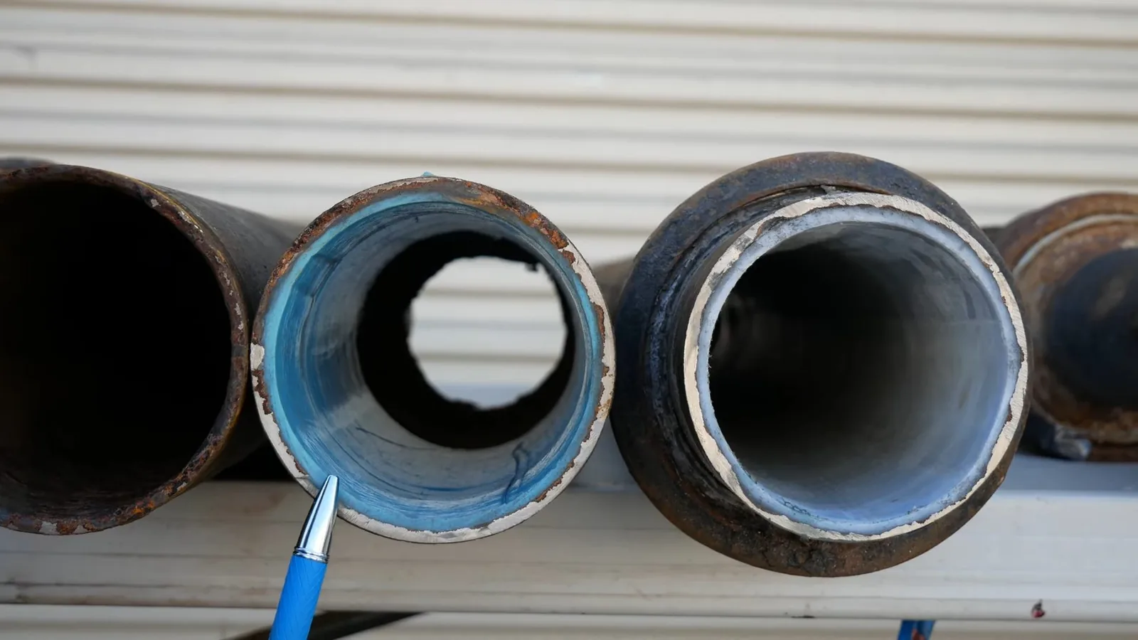 Row of cut pipe sections lined up on a ledge, one lined in blue with a pen pointing at its wall, beside rusty unlined sections