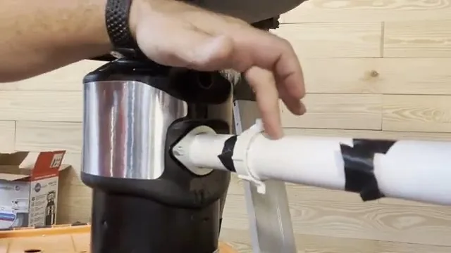 Hand pointing at the side inlet of a garbage disposal where a white pipe with black tape joins it, with a product box on the floor