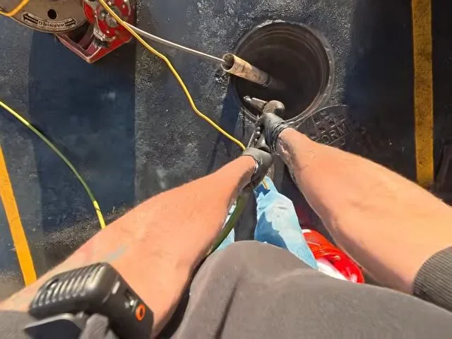 Forearms pulling a drain cable from a round cleanout opening, with a yellow cord and a two-way radio nearby