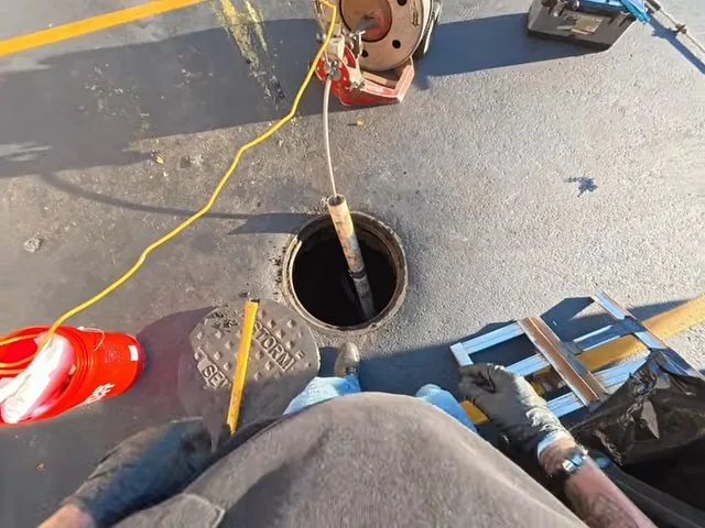 Worker's view of a drain snake being fed into an open round drain on asphalt, with a yellow cord and red equipment nearby