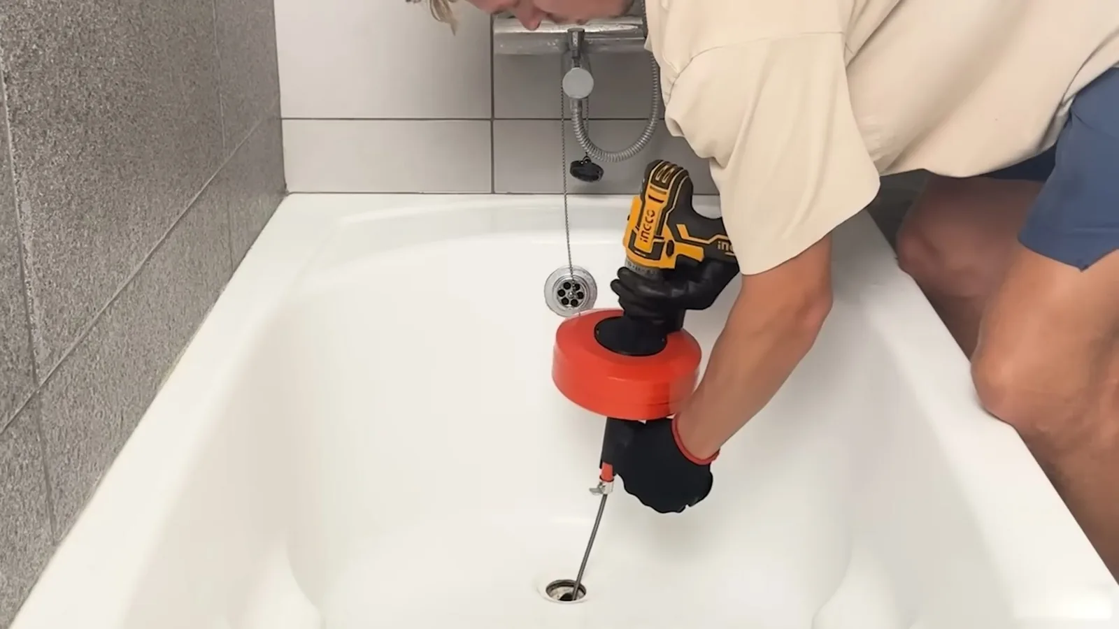 Person in a bathtub feeding a drill-powered drain auger with a red cartridge into the tub drain, one gloved hand on the cable