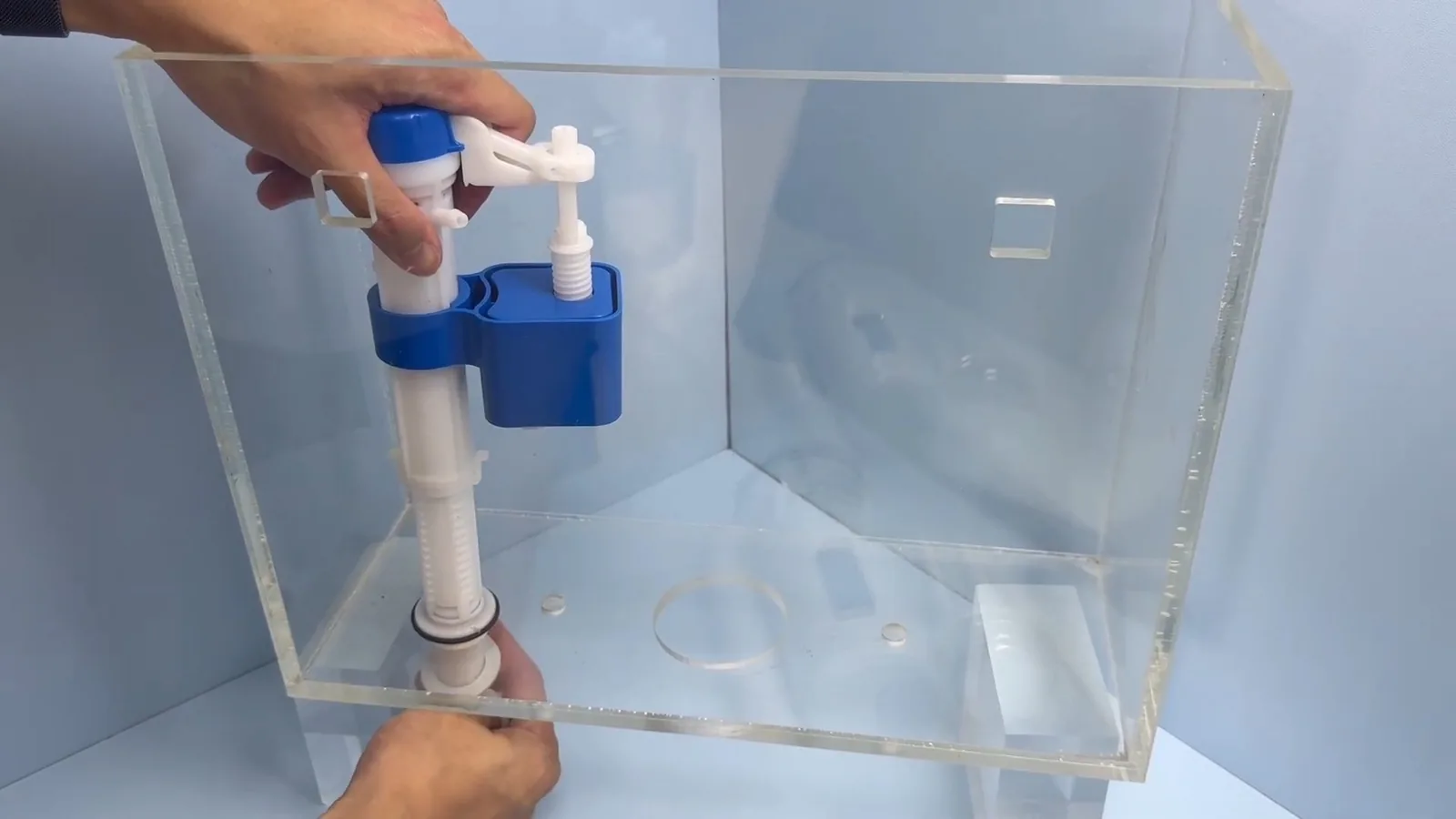 Hand placing a fill valve with a blue float inside a clear acrylic toilet tank model