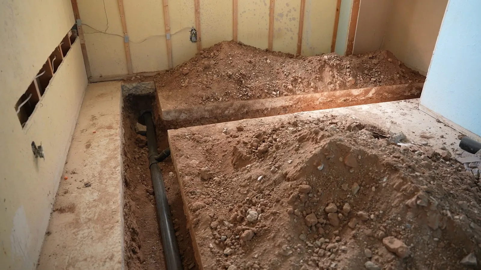 Narrow trench cut through an interior concrete floor, with a dark drain pipe visible in the soil and piles of dug earth beside it