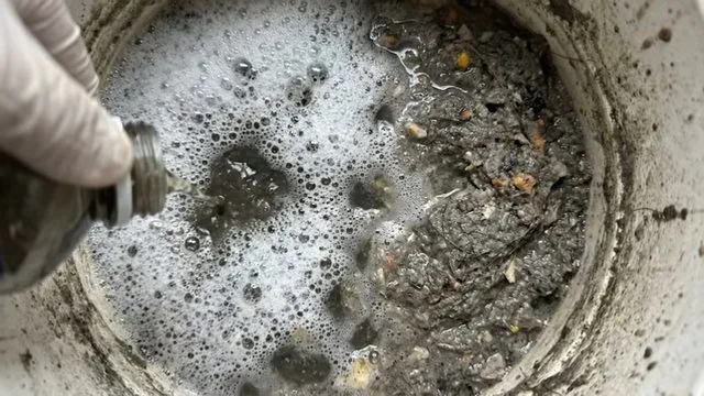 Gloved hand holding a metal fitting over a round floor drain filled with foam, silt and debris