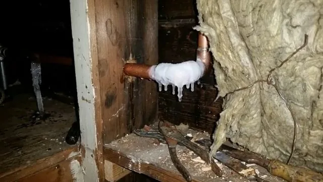 Copper elbow coated in white frost and small icicles inside a wall cavity, with torn insulation hanging beside it