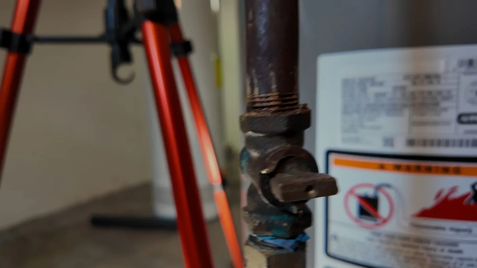 Brown gas shutoff valve with a lever handle beside a water heater label, with the orange legs of a tripod at left
