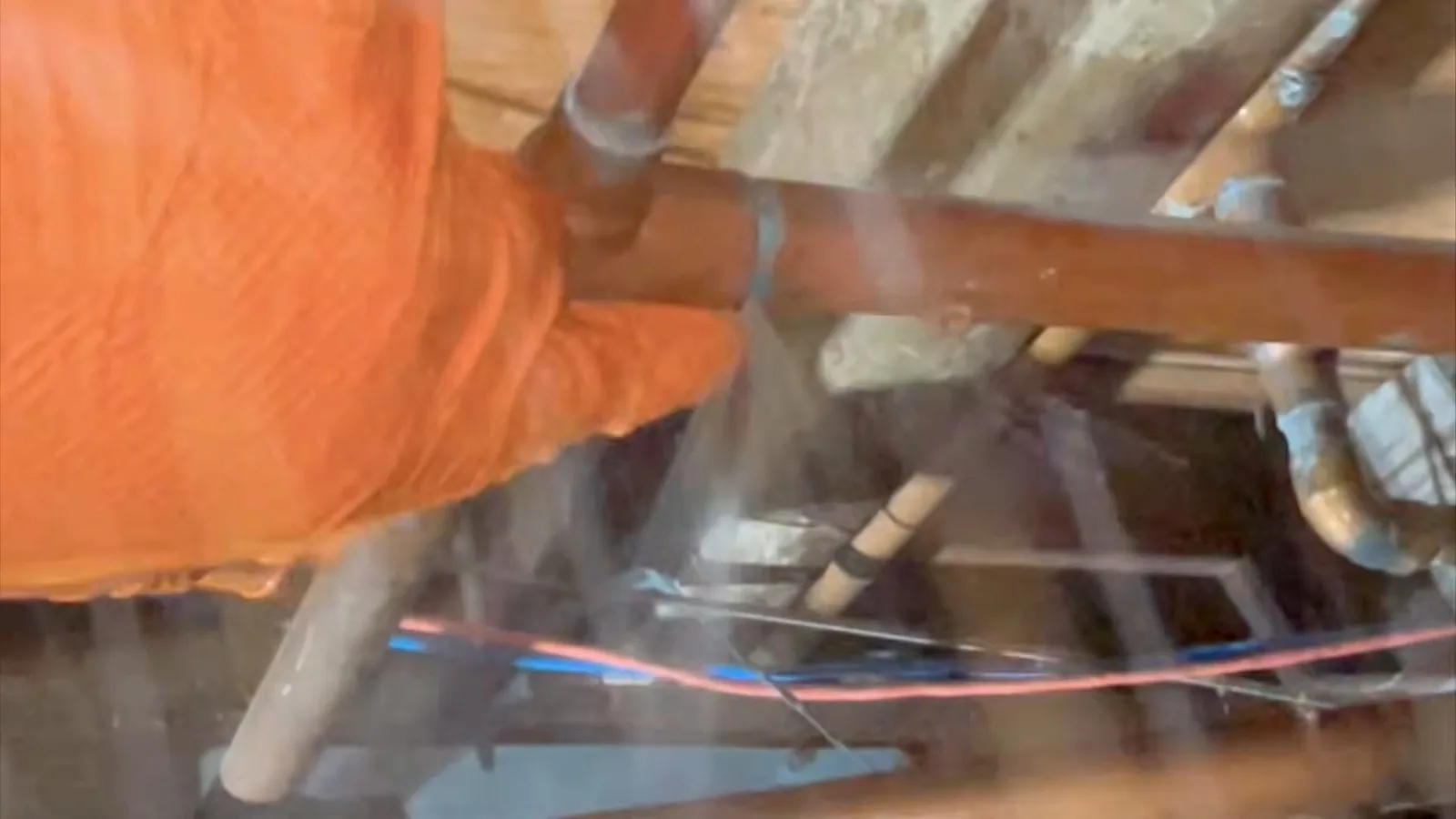 Orange-gloved hand gripping a copper pipe under floor framing while a fine spray of water comes off the pipe
