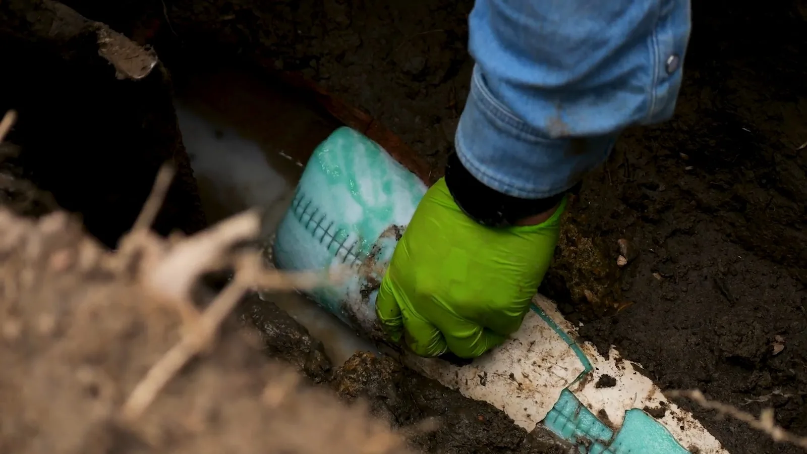 Worker in a green glove handling a pale pipe wrapped in teal material inside a dirt trench