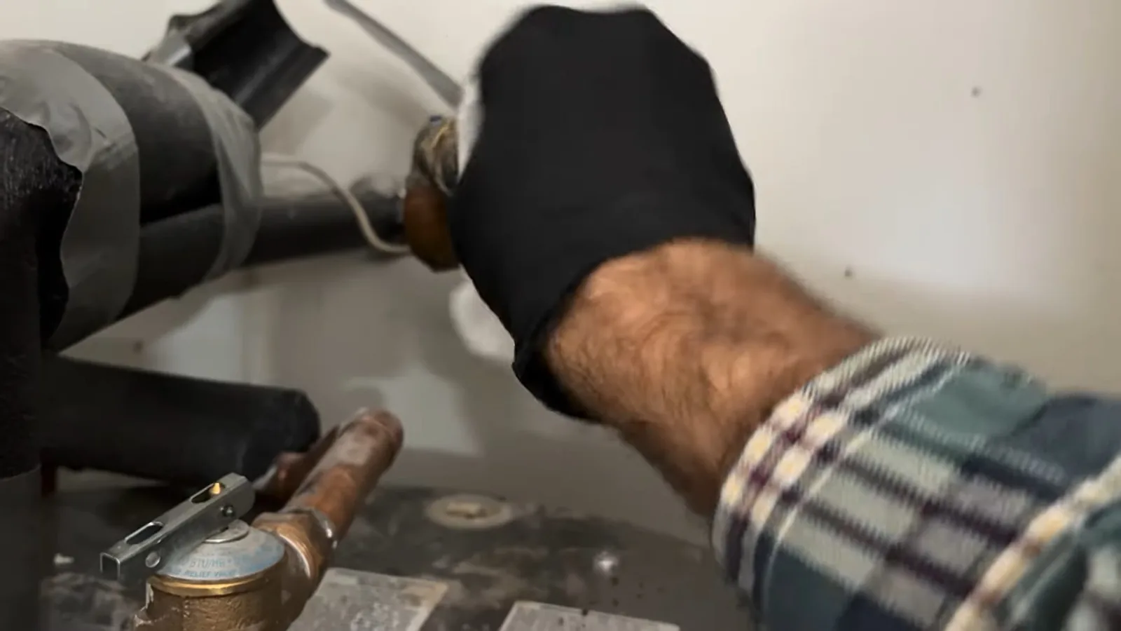 Gloved hand reaching to a pipe connection on a water heater, with a copper relief valve fitting at the bottom of the frame