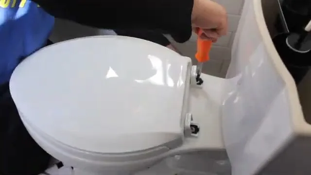 Gloved hand using an orange screwdriver on the hinge hardware of a white toilet seat