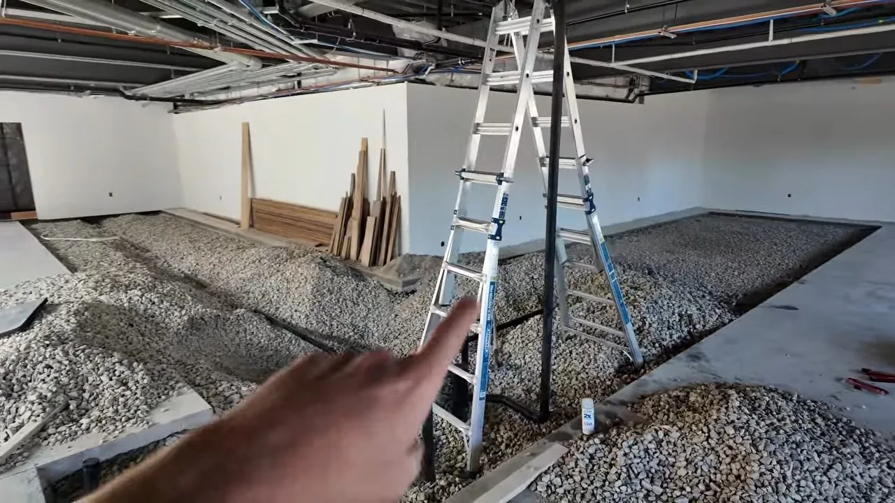 Open commercial interior with a gravel-filled trench in the floor, a stepladder, stacked lumber and a hand pointing toward the ladder