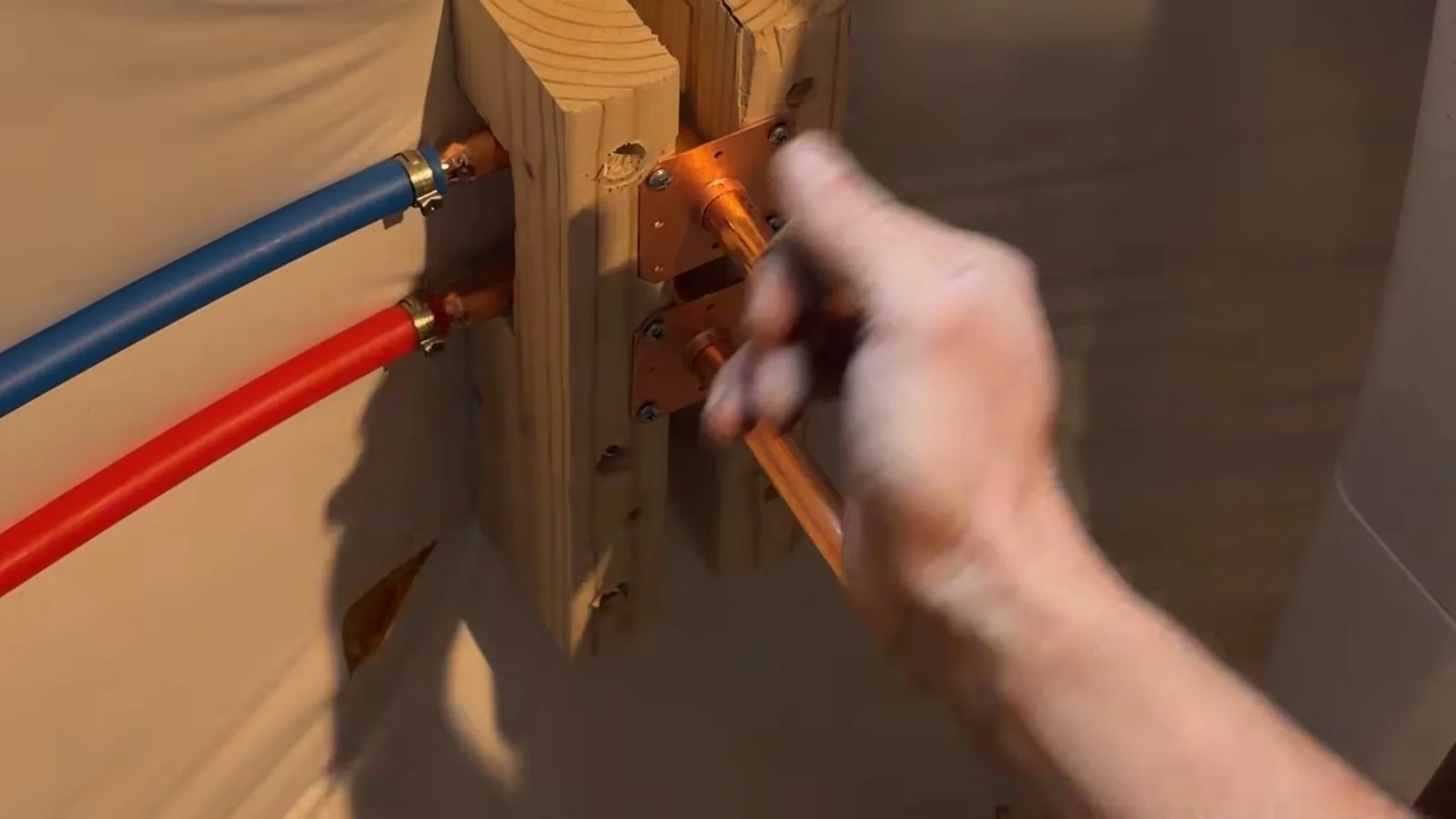 Hand working at copper fittings where blue and red flexible supply lines attach to a wood mounting board