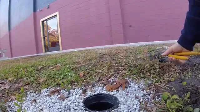 Hand guiding a yellow cable into a ground-level cleanout surrounded by gravel, with a pink-walled building behind