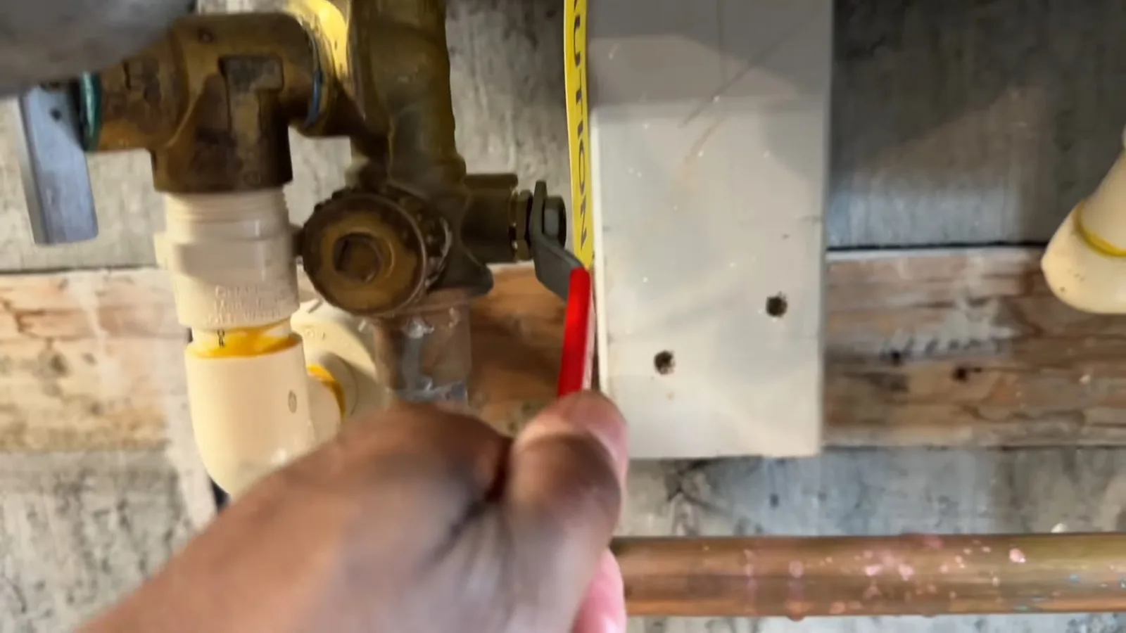 Hand holding a tape measure across brass valves and white pipe fittings beside a pale panel with two drilled holes, with copper pipe below