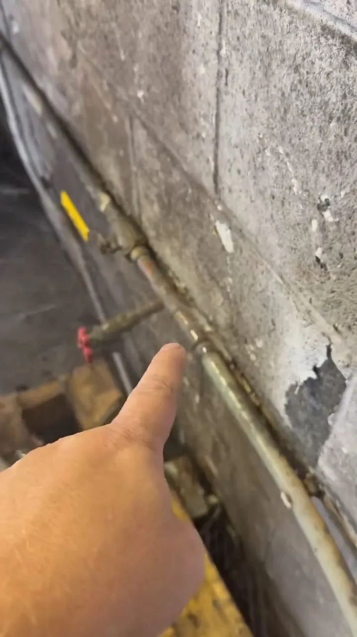 Hand pointing to a copper pipe running along a painted concrete block wall with flaking paint
