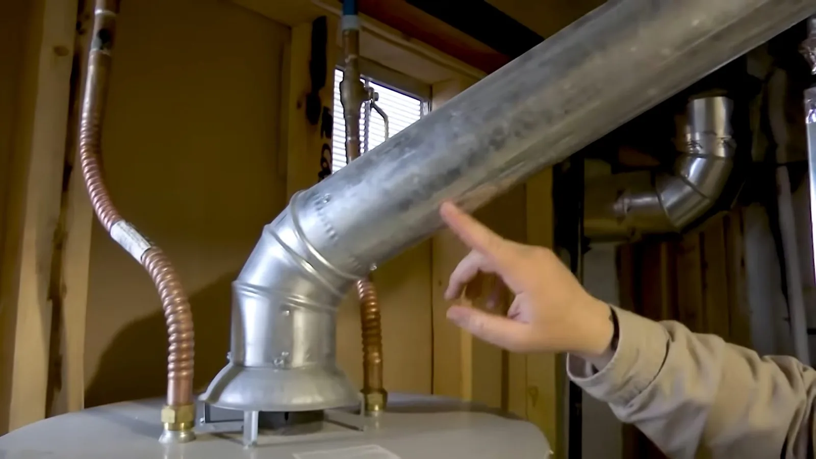 Hand pointing at a galvanized vent elbow where it meets the top of a water heater, with a copper gas line and a window behind