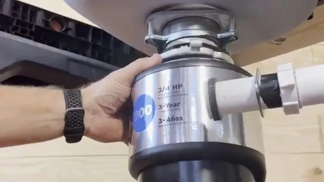Hand steadying a stainless garbage disposal mounted under a sink, with a white discharge pipe attached at the right