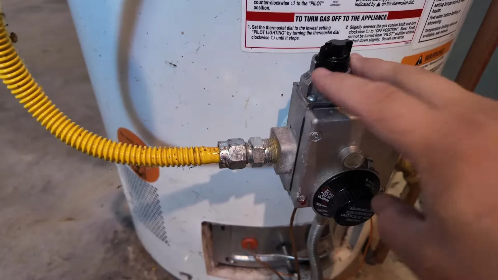 Hand turning the dial on a gas water heater control, with a yellow flexible gas line and a lighting-instructions label above