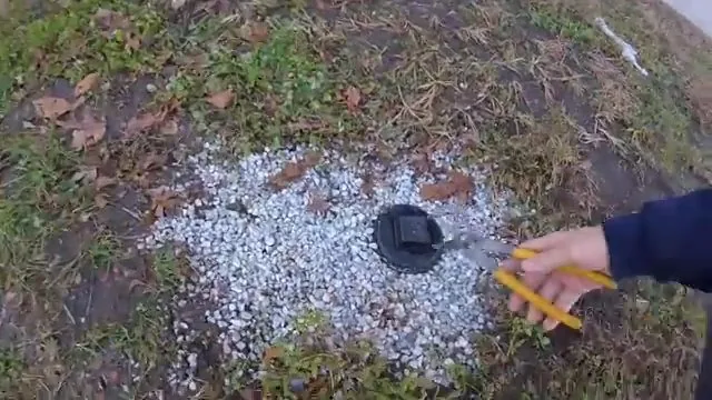Hand holding a yellow pencil near a black cleanout cap set in a patch of white gravel in grass