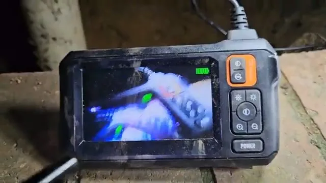 Handheld inspection camera monitor showing a blurry blue and green pipe interior, resting on a rough surface