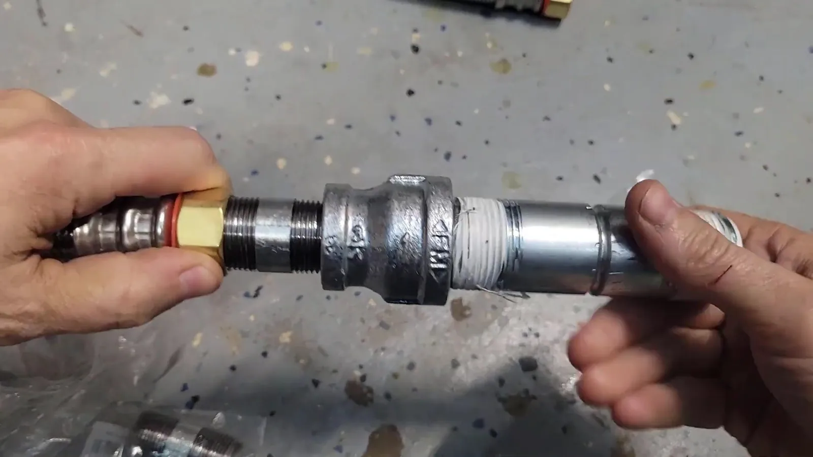 Two hands joining a galvanized coupling between a brass fitting and a steel pipe, with white thread tape on one thread