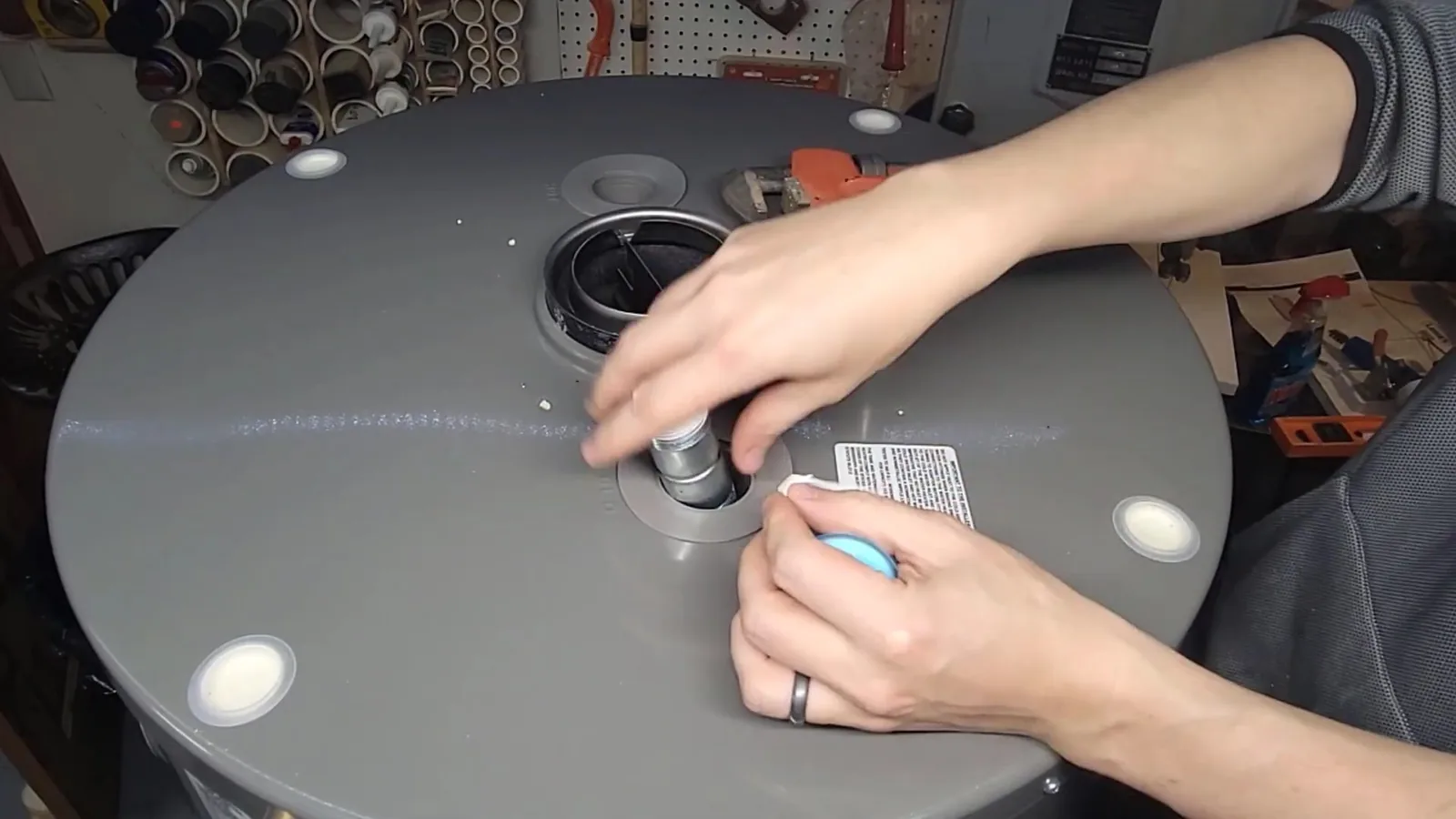 Hands working on a fitting on top of a gray water heater tank in a workshop with pegboard behind