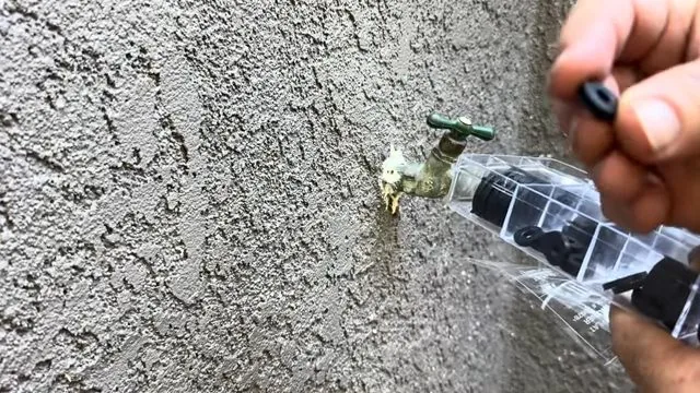 Brass hose bib with a green handle on a gray stucco wall, with a hand holding a small black part and a clear package nearby