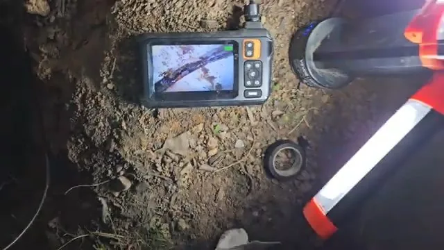Handheld camera monitor lying on dirt beside a cut pipe in an excavation, with an orange work light at the lower right