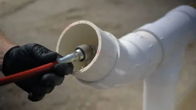 Gloved hand feeding a metal jetting nozzle on a red hose into the open end of a white PVC pipe fitting
