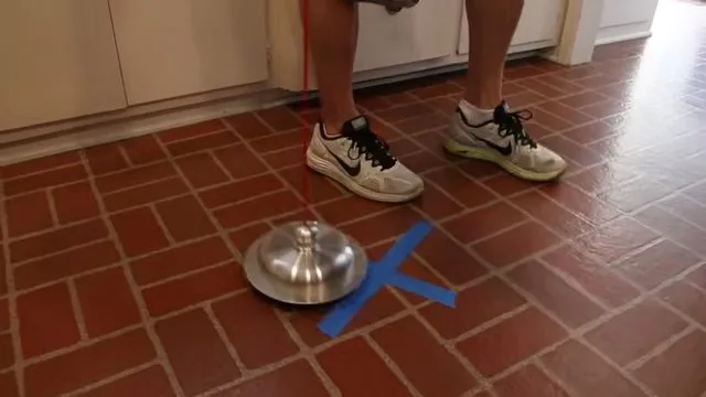 Metal listening disc resting on a red tile kitchen floor beside a blue tape cross, with a person standing nearby