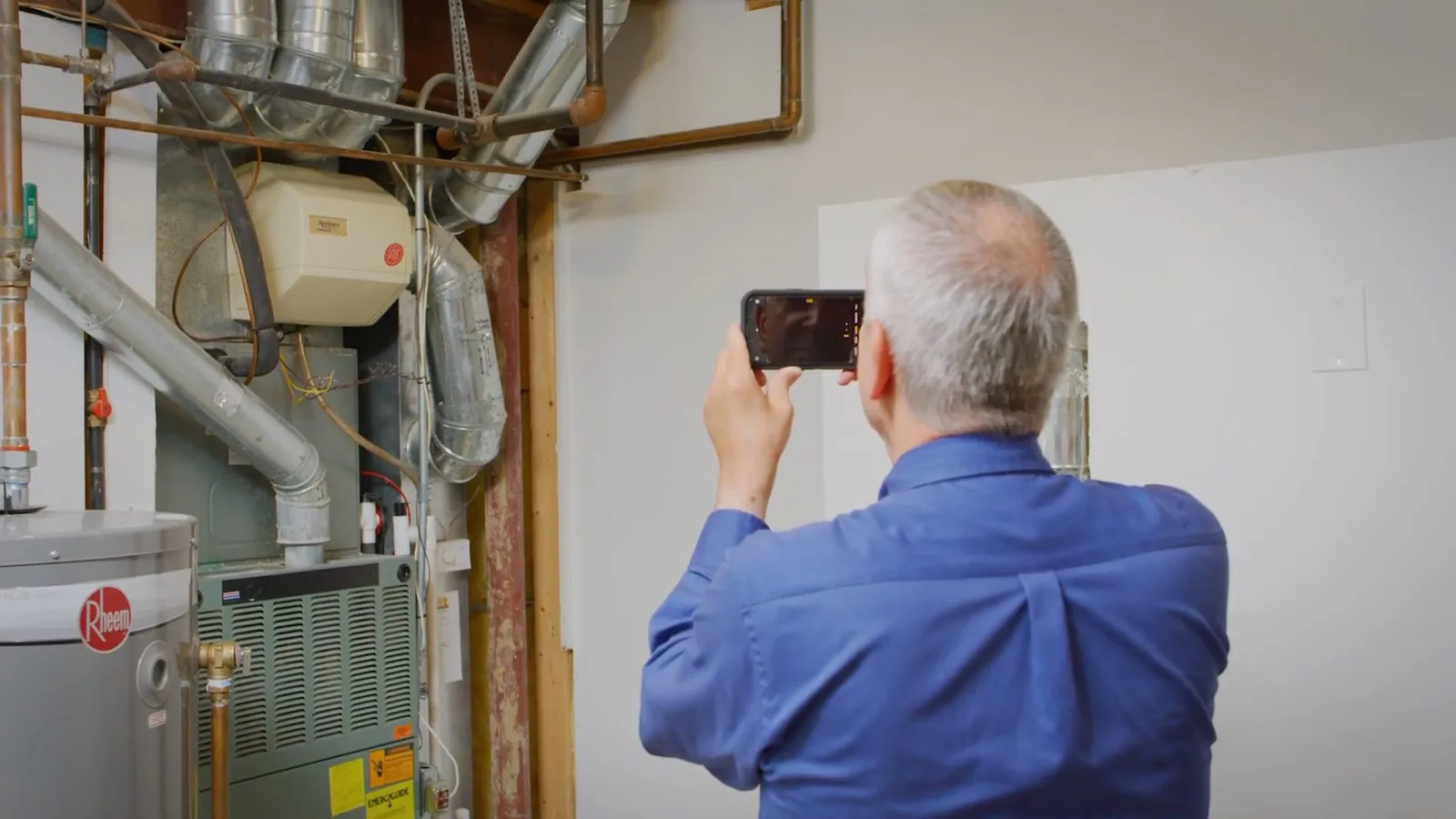 Man in a blue shirt holding a phone up to photograph a water heater and furnace in a utility room with exposed pipes and vent