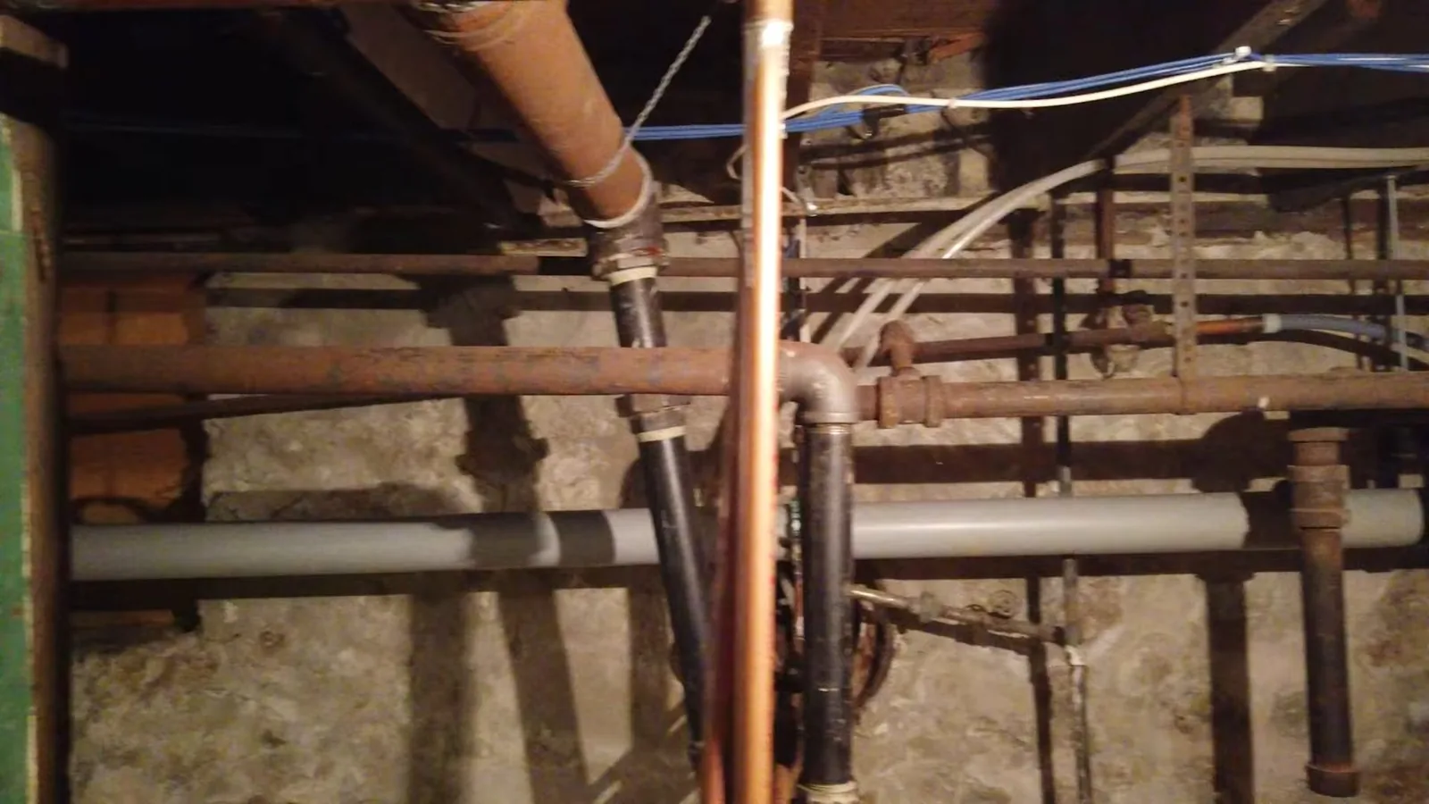 Several different pipes of copper and other materials running along a basement wall beneath floor joists