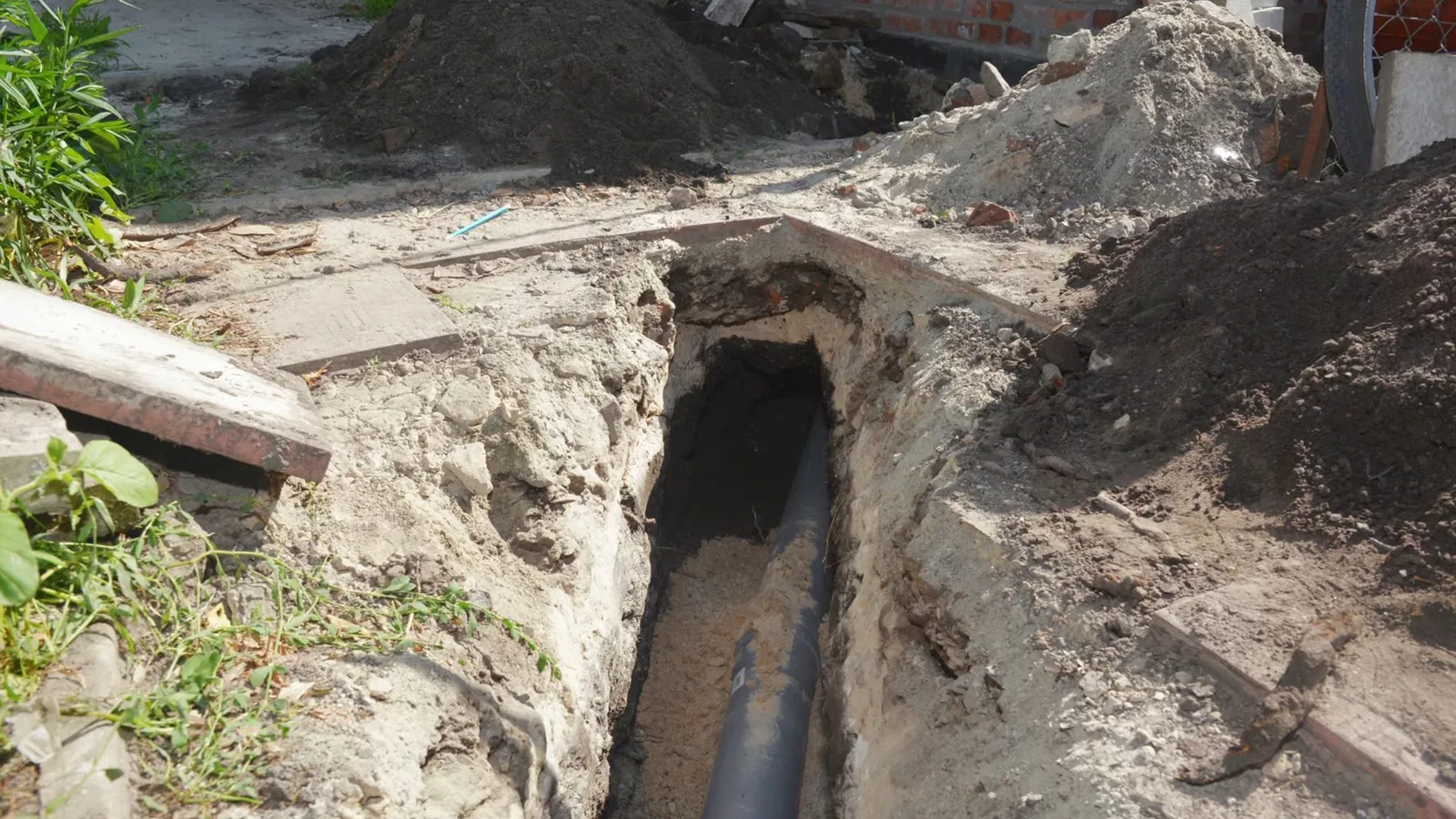 Narrow trench dug beside a house wall with a dark pipe visible in the bottom and soil piled at the sides