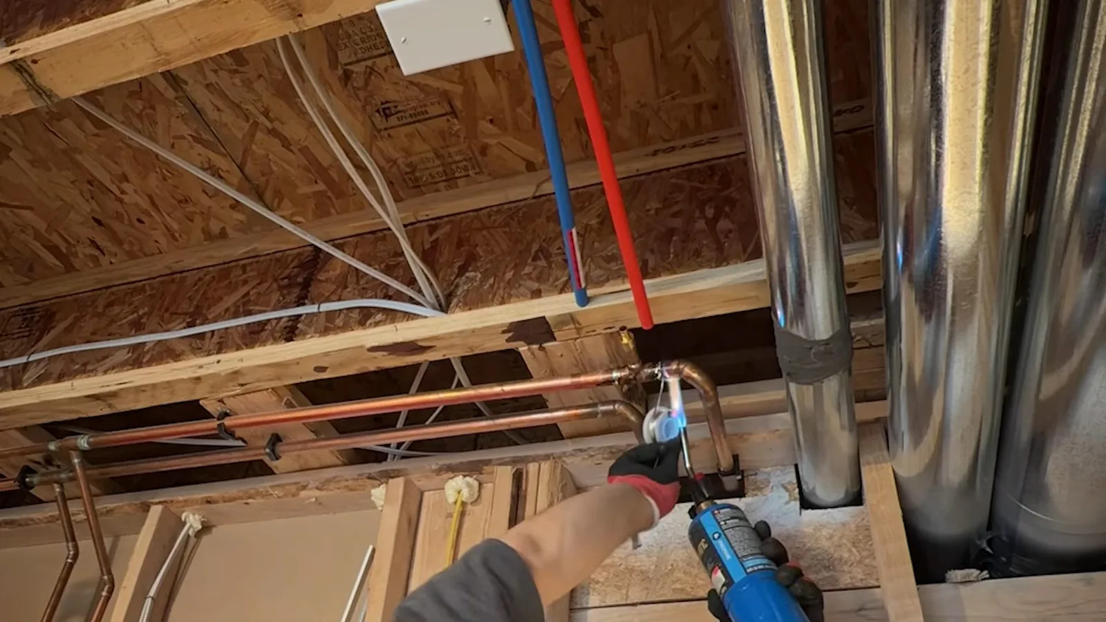 Open basement ceiling with new copper pipe and red and blue flexible lines run between joists, and a plumber soldering a joint
