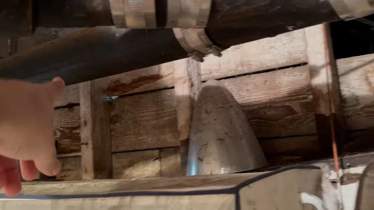 Hand pointing toward an old cast iron drain pipe with clamps below a wooden subfloor and joists, with rust marks on the wood