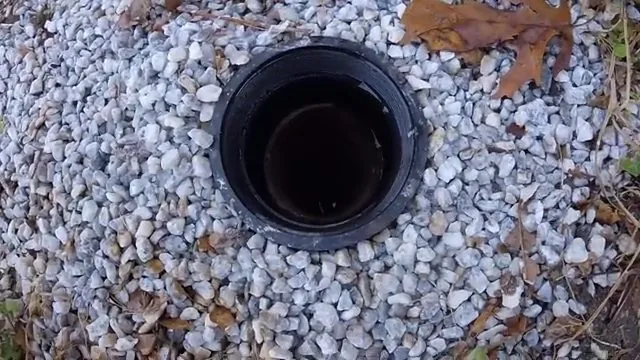 Open black sewer cleanout fitting set in a bed of gray gravel with fallen leaves around it