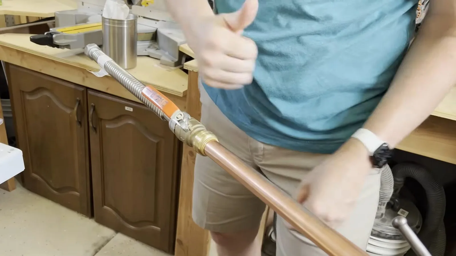 Person in a blue shirt giving a thumbs up while holding a flexible metal connector joined to a copper pipe, with wooden cabinets and a workbench behind