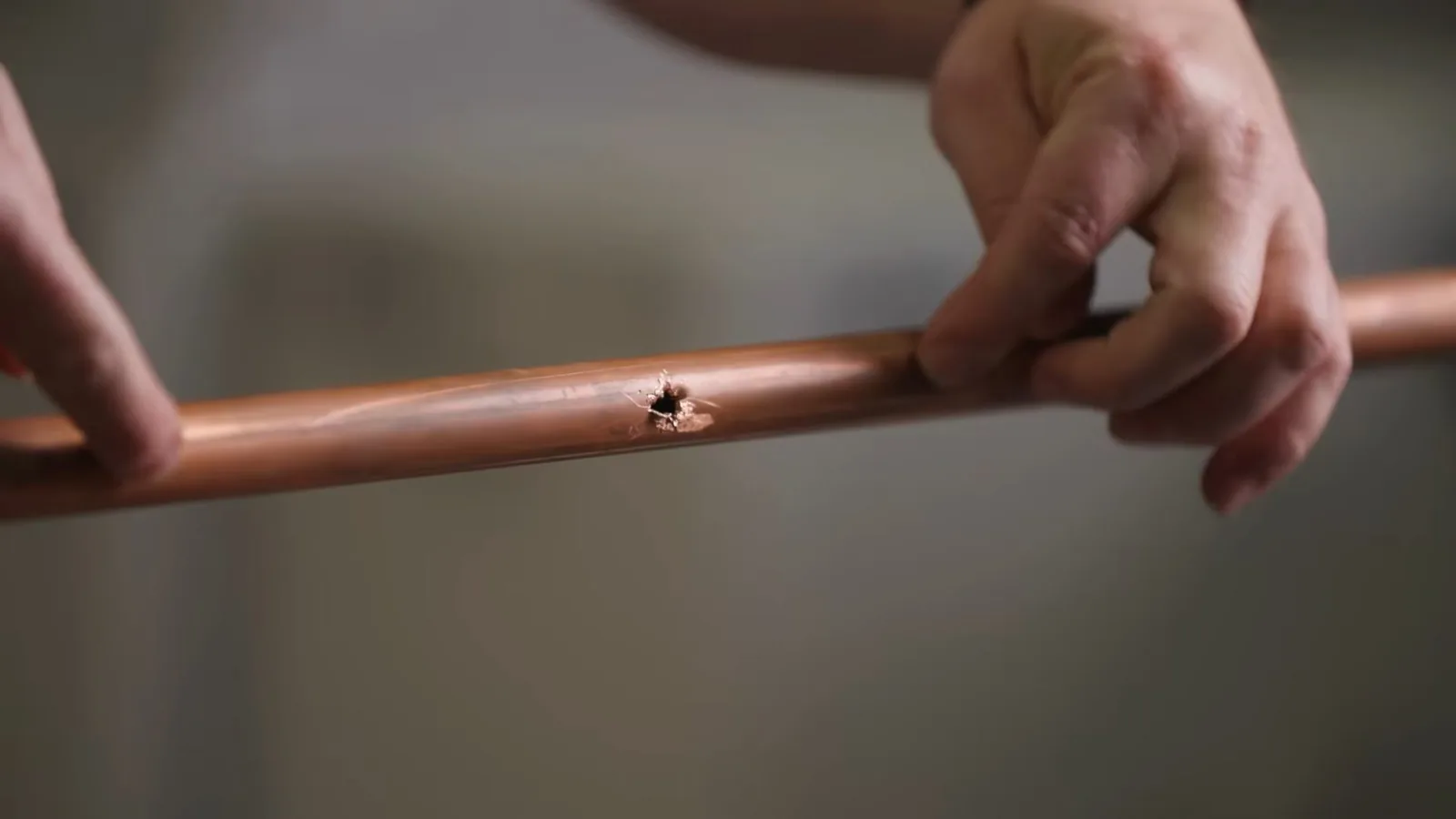Two hands holding a length of copper pipe with a small pinhole in the side