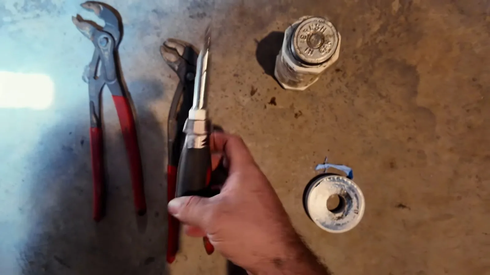 Hand holding a screwdriver beside slip-joint pliers, a pipe wrench, a capped brass fitting and a roll of thread tape on a concrete floor