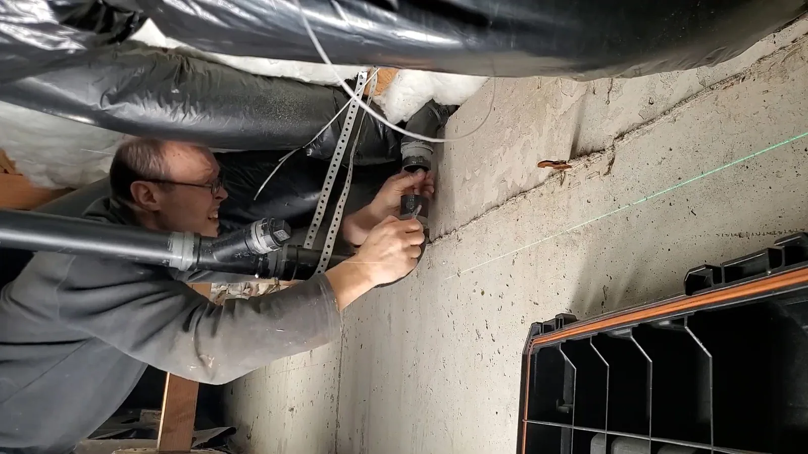 Smiling man in a gray shirt reaching overhead between floor joists to work on a pipe fitting, with black sheeting and insulation above him